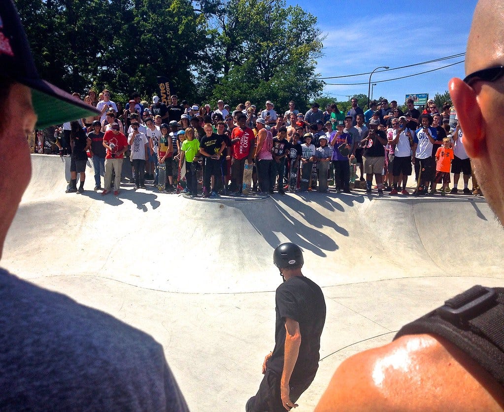 A pair of people’s shoulders in the foreground frame Tony Hawk in a skatepark bowl skating toward a crowd of children watching him on the opposite lip.