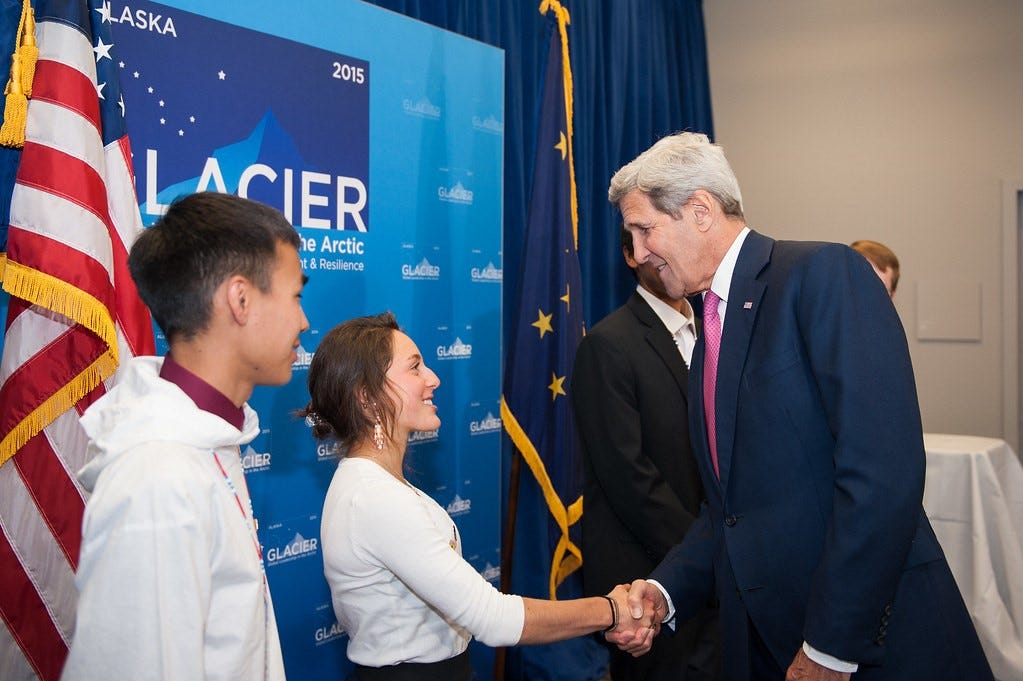 young woman shakes an older man’s hand in front of a sign that reads glacier and an american flag