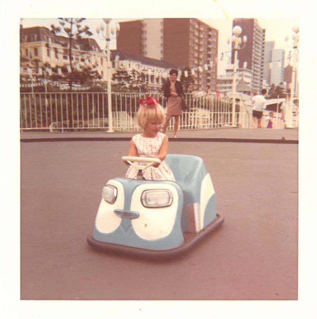 A young female child in a blue bumper car with her mother and some buildings in the background