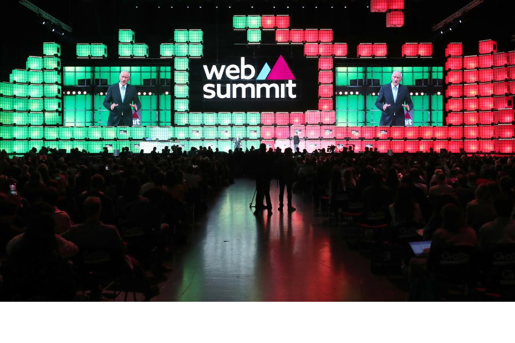 Imagem distante do palco principal do evento, iluminado com as cores vermelho e verde representando as cores da bandeira de Portugal. O presidente de Portugal Marcelo Rebelo de Sousa aparece em destaque nos telões do palco principal do evento, ele usa um terno preto com camisa azul.