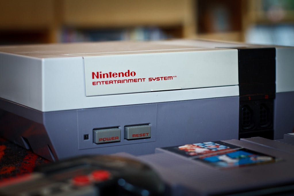 A close-up photo of a Nintendo Entertainment System console. A blurry Super Mario Bros. cartridge is in the bottom right, and some even blurrier cables (completely out of focus) are in the bottom left.