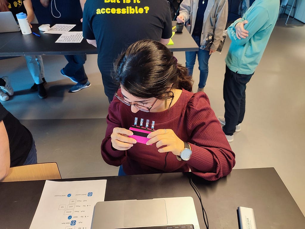 A woman with dark hair in a ponytail is sitting in front of a laptop and holding a pink 3D-printed sip & puff device whilst reading a sheet of commands.