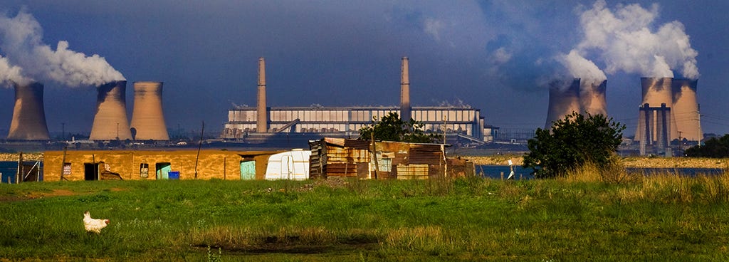 Green grass and a chicken in the foreground, dilapidated buildings in the middle, and smoke stacks spewing grey into the blew sky in the background.
