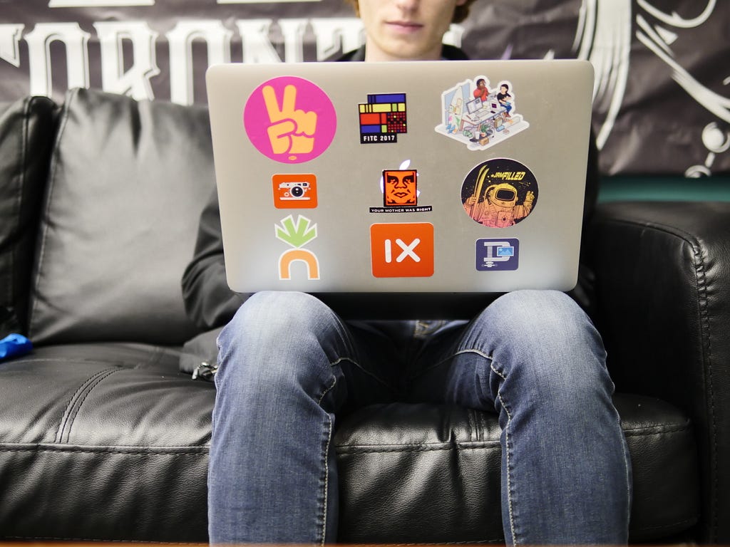 A person wearing jeans sitting on a black leather sofa with a laptop that has stickers on the laptop lid, including one that covers the Apple logo. This indicates that this person has adopted the laptop by personalizing it and is using it in regular life.