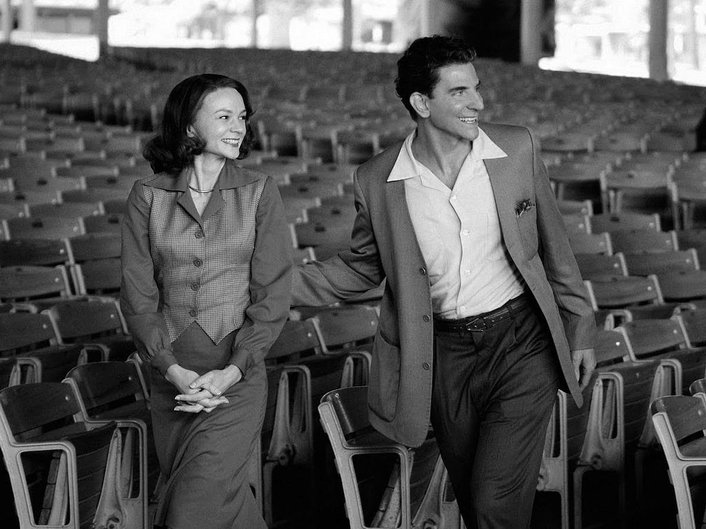 A woman and a man smile while walking through an auditorium. (Carey Mulligan playing Felicia Montelegre and Bradley Cooper playing Leonard Bernstein)