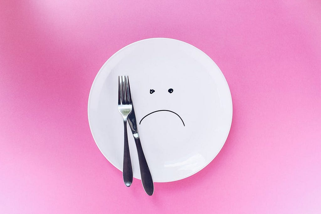 A fork and a knife sitting on the edge of a white plate. A sad face is drawn on the white plate.