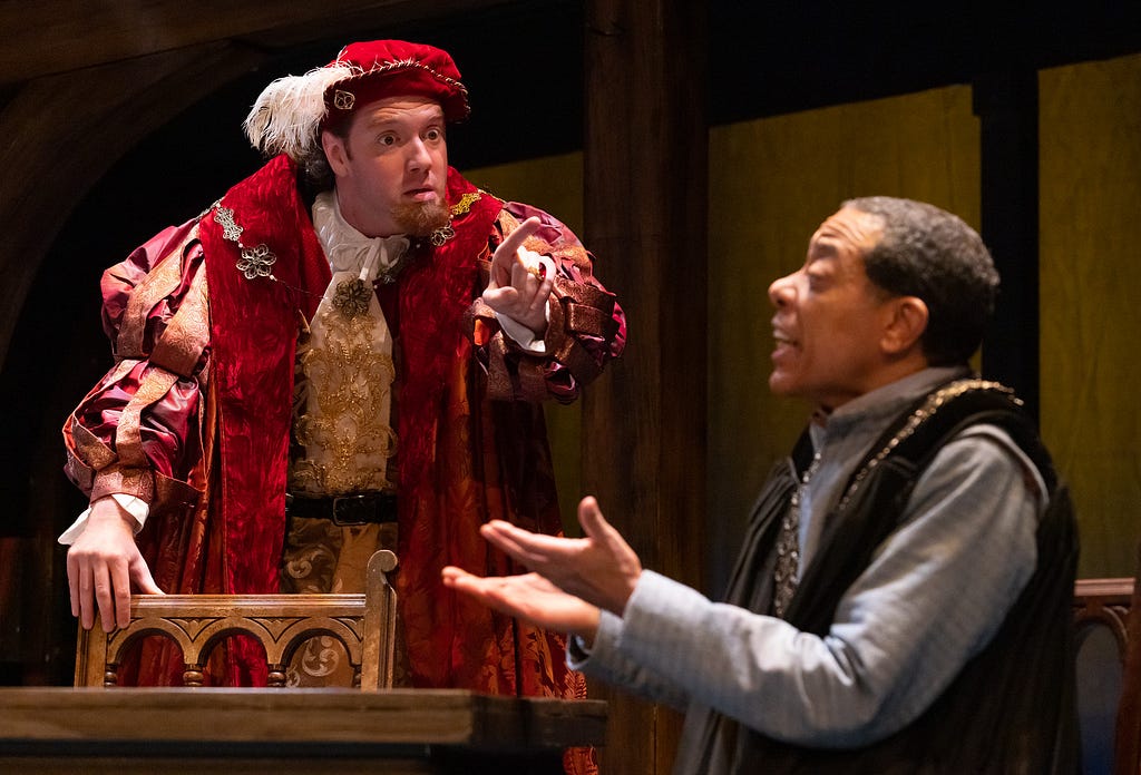 A man with light skin in extravagant red and gold 16th century clothes and hat points at a man with brown skin and hair in a simple blue-gray shirt and black robe.