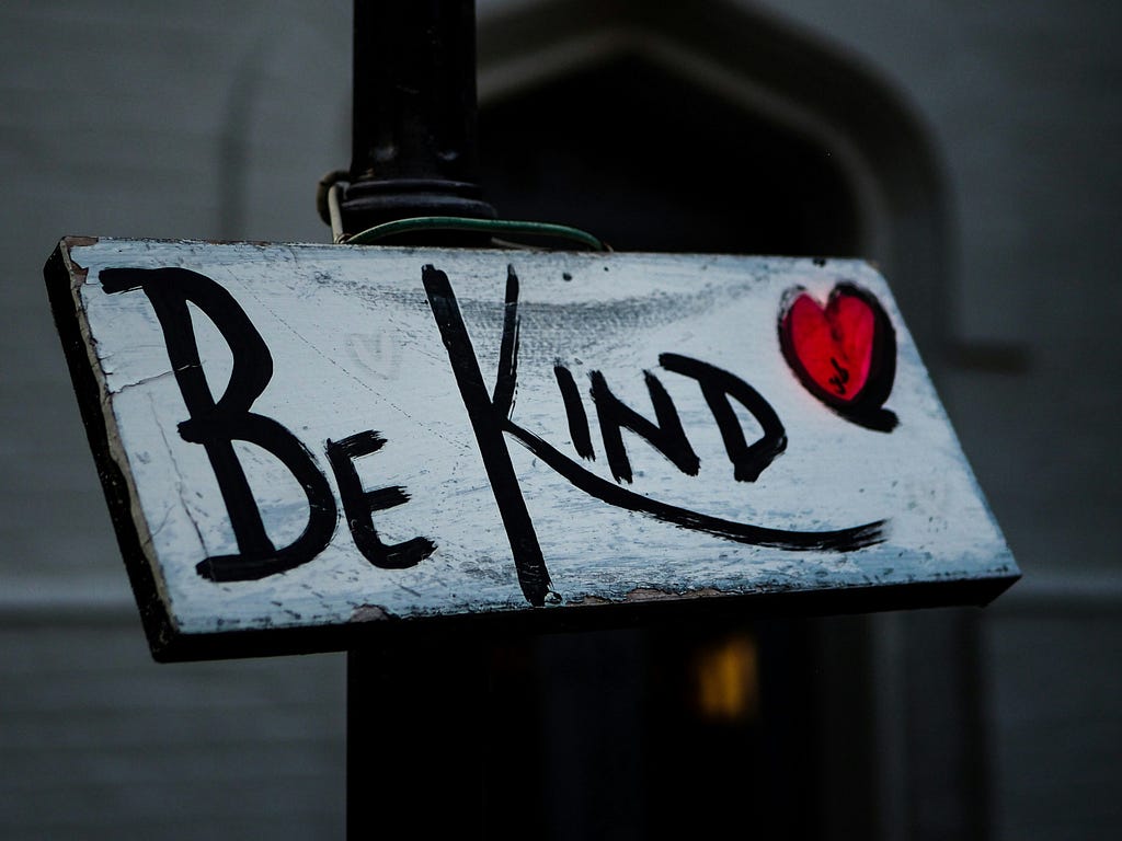 Photo of a wooden sign on a light post, the sign has a white painted background and in black letters it reads “Be Kind” next to a red heart.