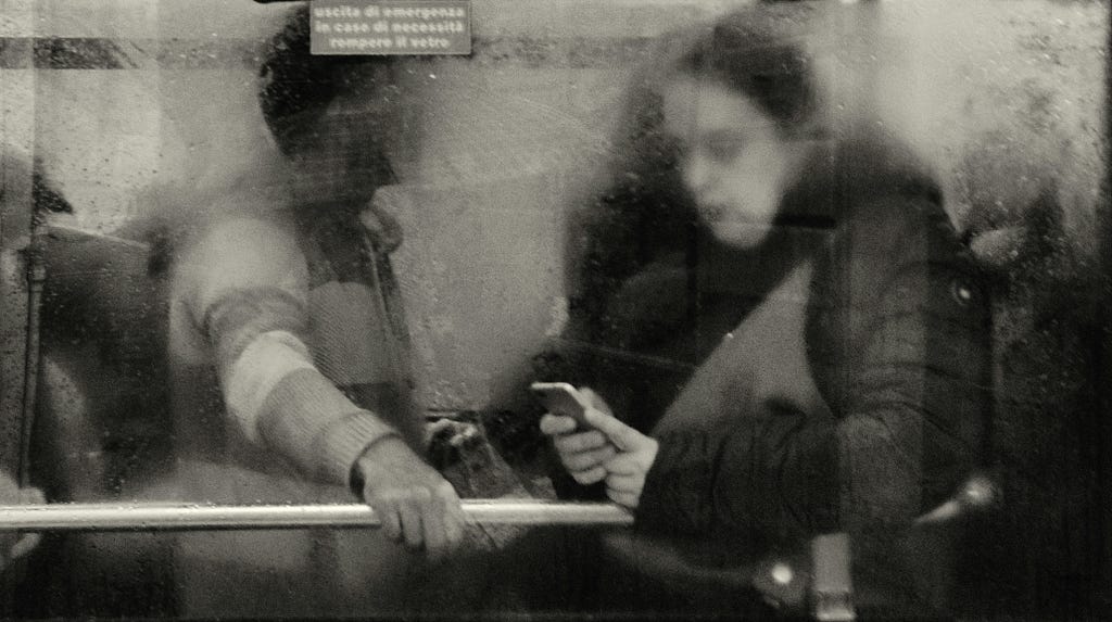 Two people seen through a fogged and dirty glass window; one leans on a railing while the other looks down at a phone, absorbed in the screen.