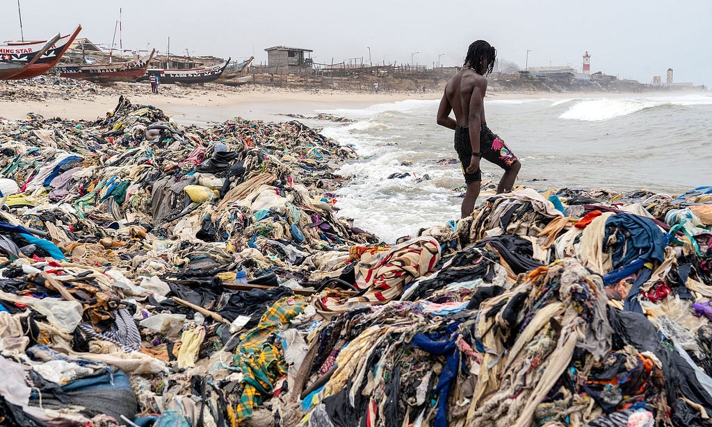 a person standing atop a heaping pile of clothes on a beach in Ghana