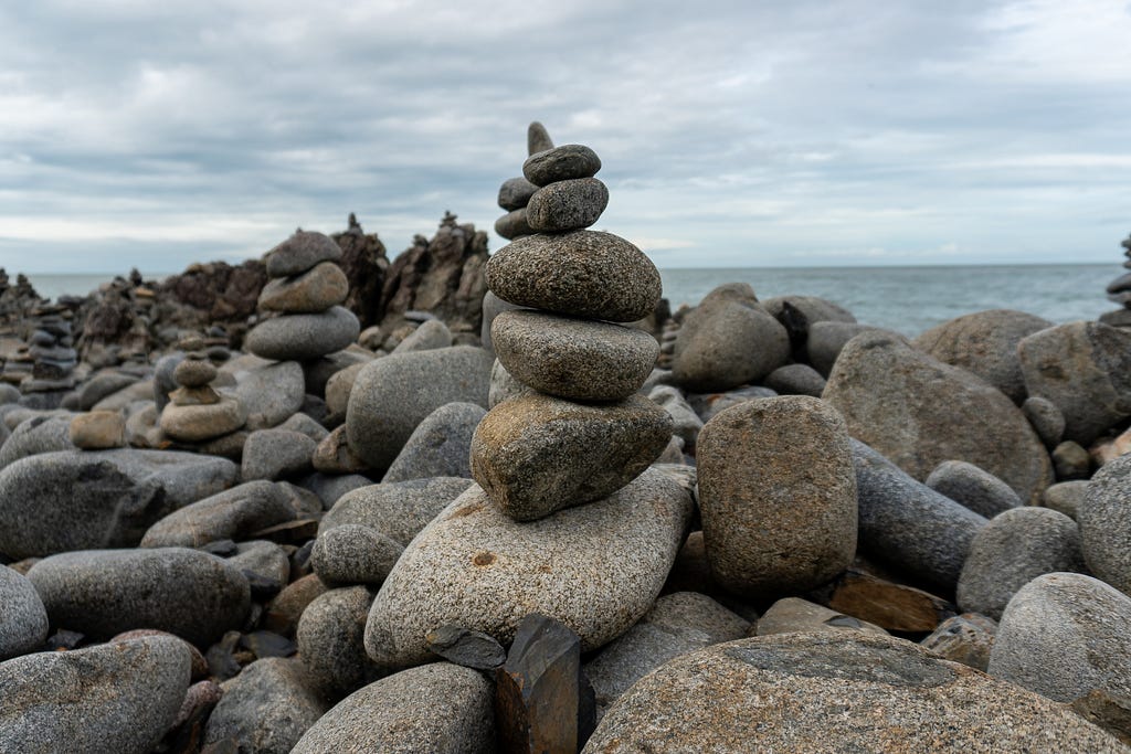 Pequenas pedras equilibradas em paisagem com diversas pedras representando "equilíbrio" com o mar ao fundo.