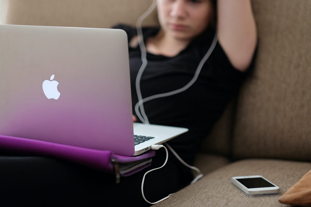 Girl sitting on a sofa, with earphone plugged into a laptop.