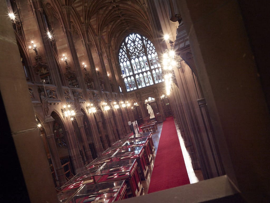 A long view of the historic reading room at the John Rylands Library featuring neo-gothic architecture and stained glass window.