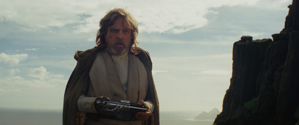 Luke Skywalker holds his lightsaber, which Rey has just given him. He is about to throw it over his shoulder and begin the process of upending our expectations.