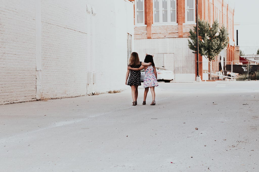Two young girls, kids, walking down the street, arms about each other.