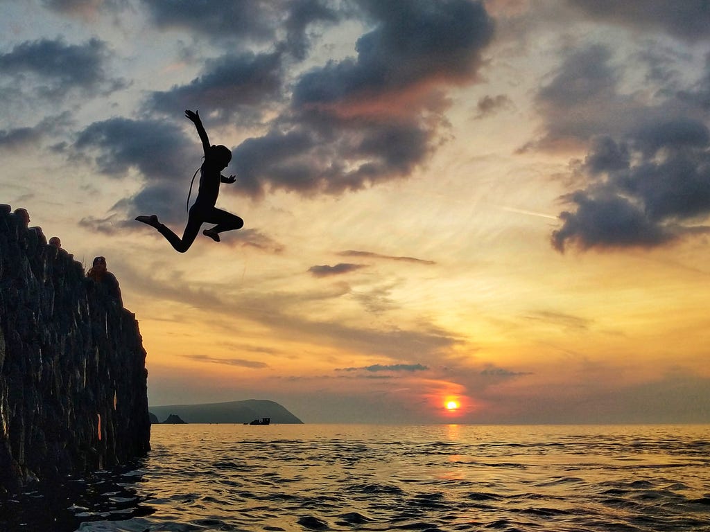 A person, silhouetted by sunset, jumps into the ocean from a cliff.