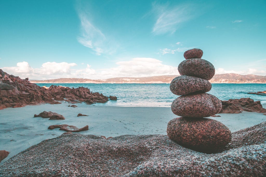 A tower of rocks called a cairn balances overlooking a sunny beach. Each rock contributes to its ability to stand tall.