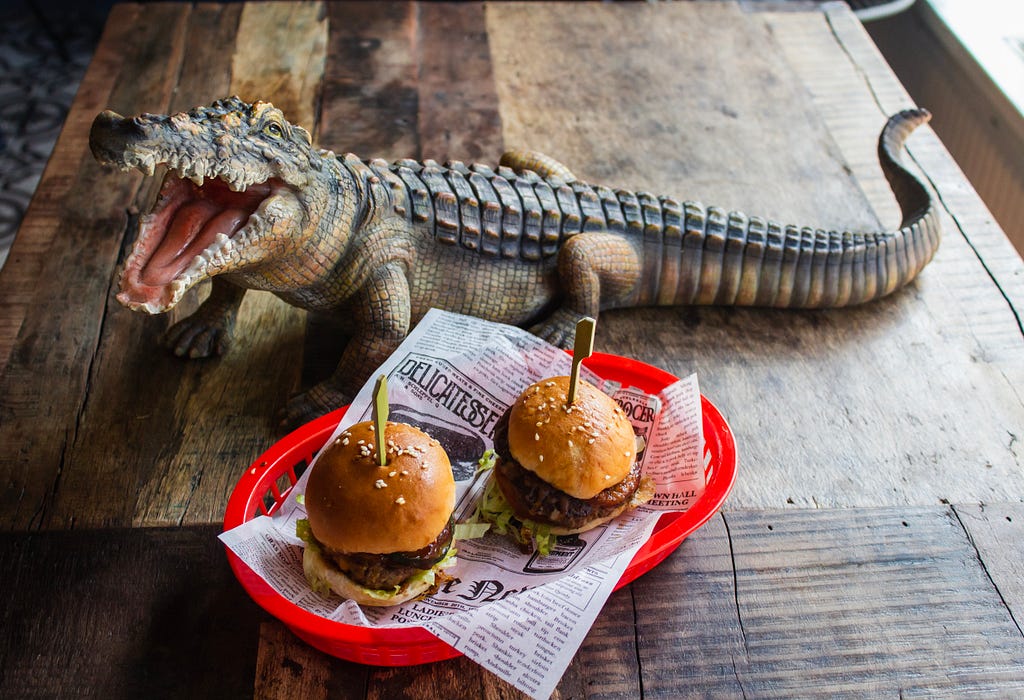 A couple of fresh burgers served in a red basket at a Louisiana bar. An alligator statue sits on the table with the burgers.