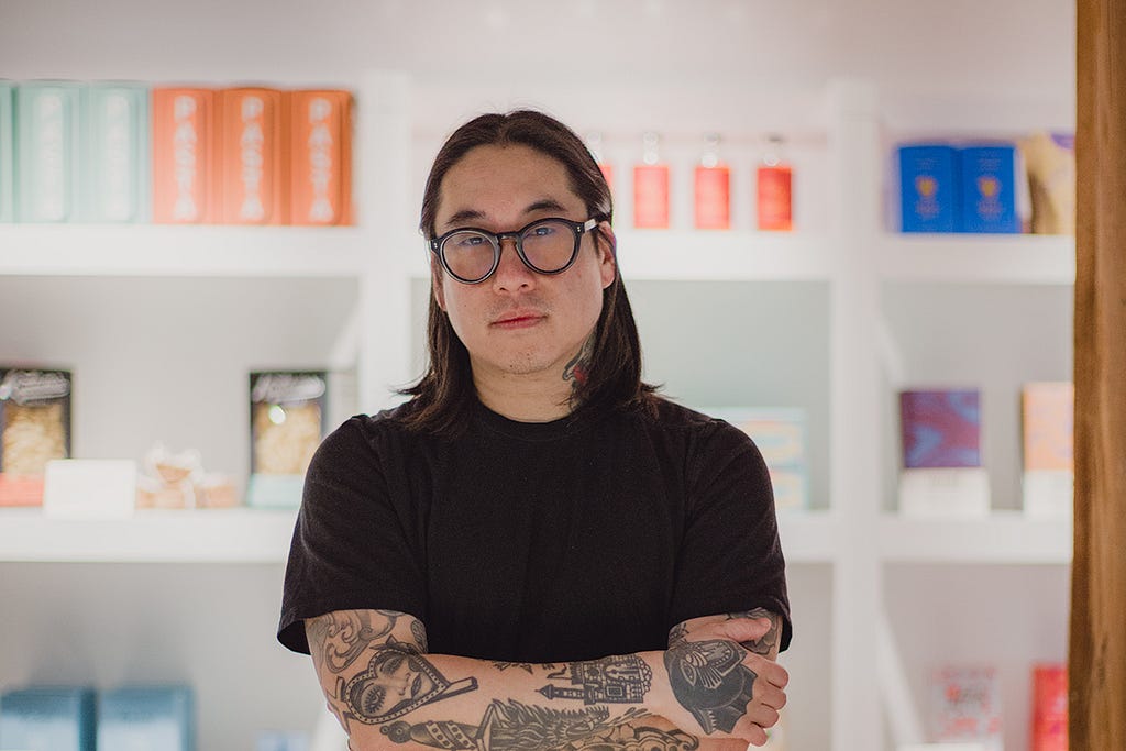 Ryan Lee headshot. Ryan is wearing a black shirt with his arms crossed over his chest. He has dark, shoulder-length hair and is wearing thick-rimmed glasses.