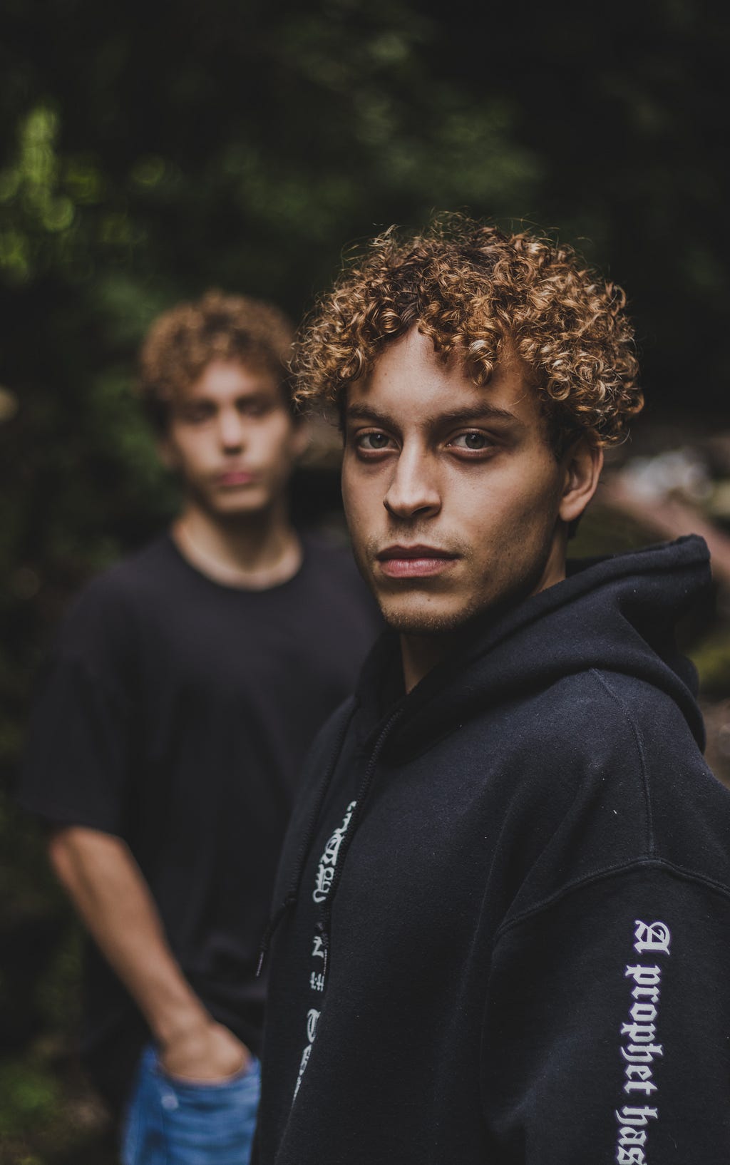 A male-presenting person with curly hair looks into the camera in the foreground while an identical person is blurred in the background.