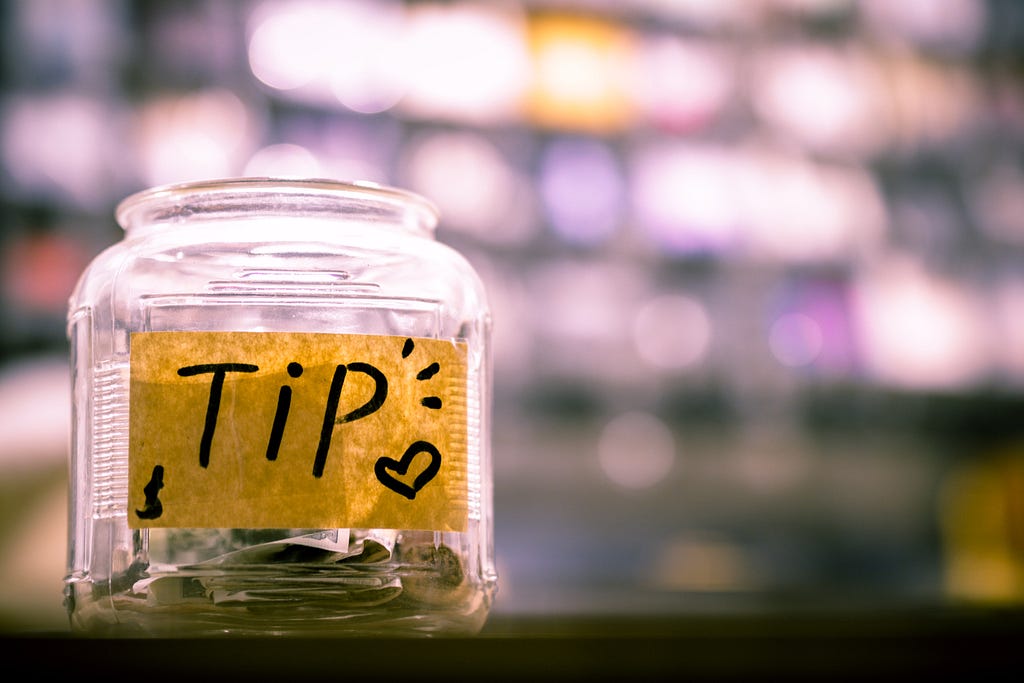 A clear, transparent jar with a yellow sticker saying “Tips” on it. The jar has some coins in it.