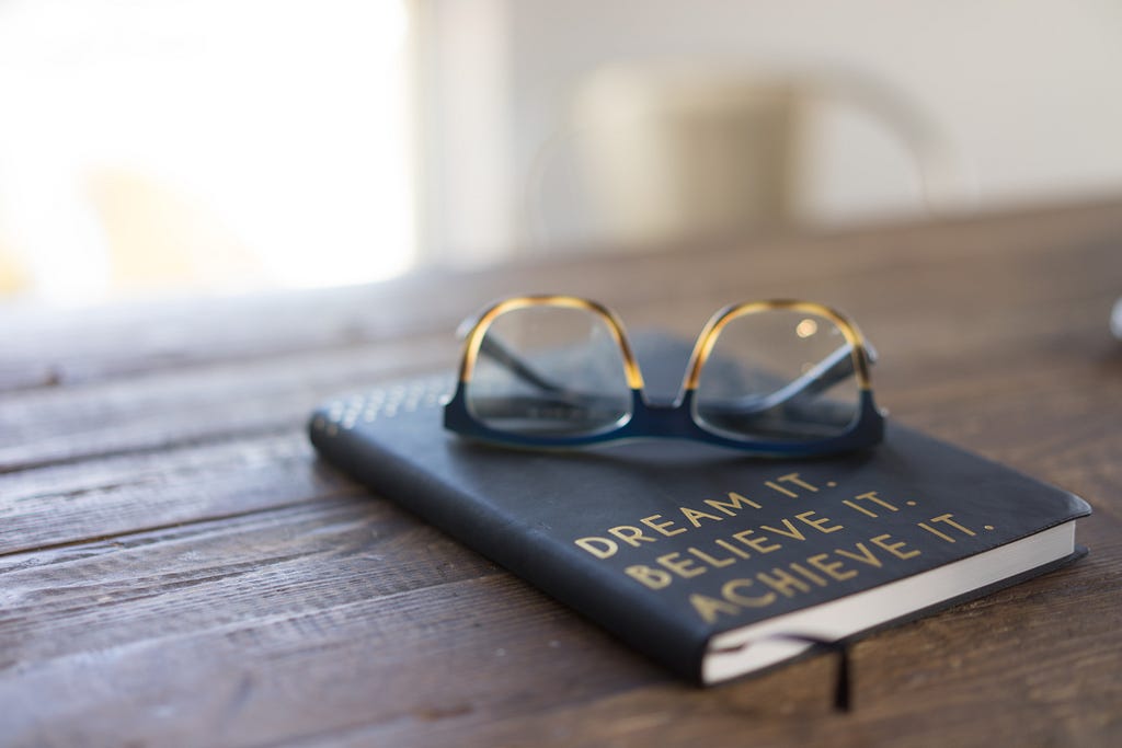 A pair of glasses and journal on table with text “Dream it, Believe it, Achieve it” on cover.