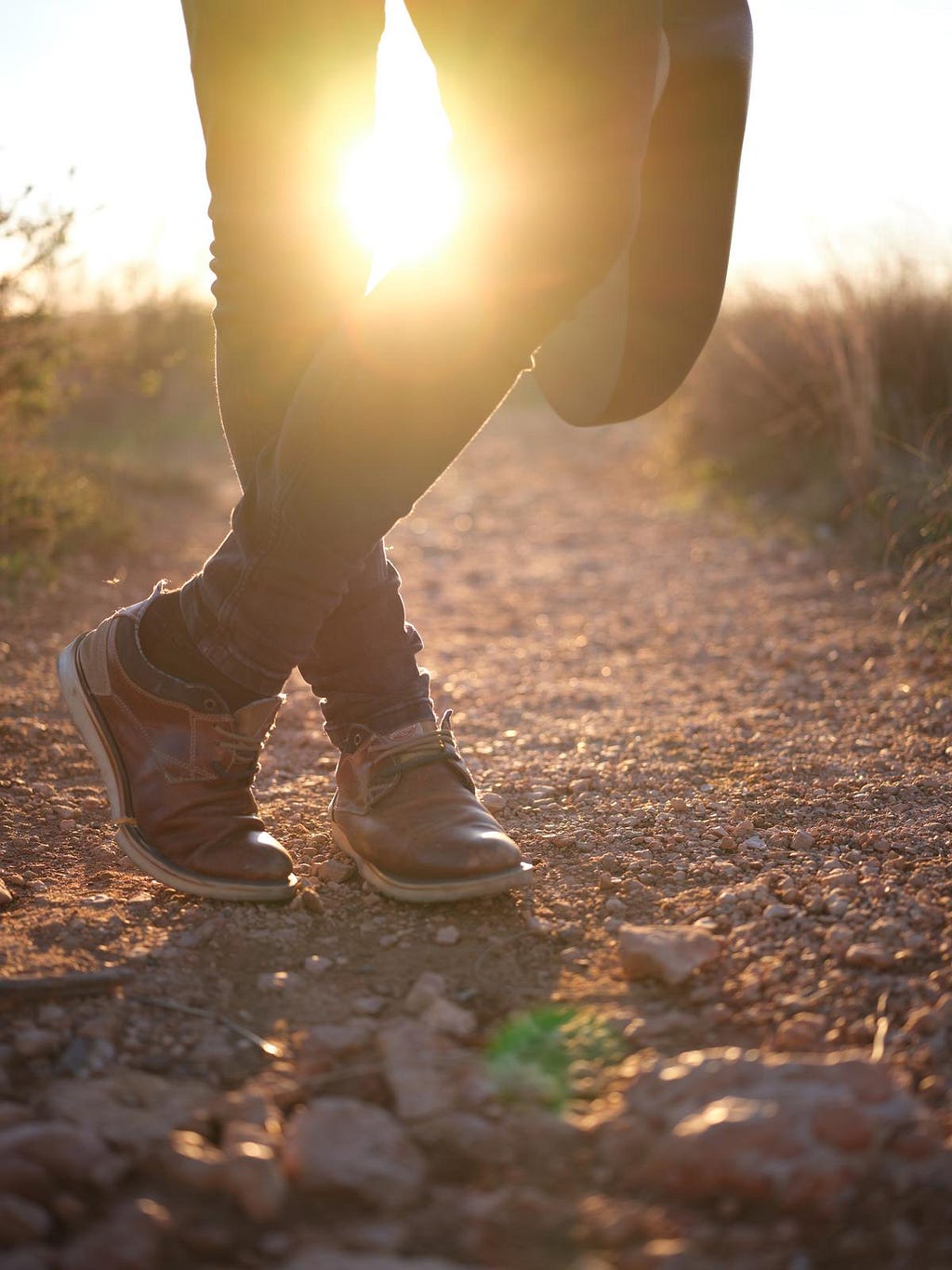 Person standing with ankles crossed on a gravel road. Sunlight shines between their knees.