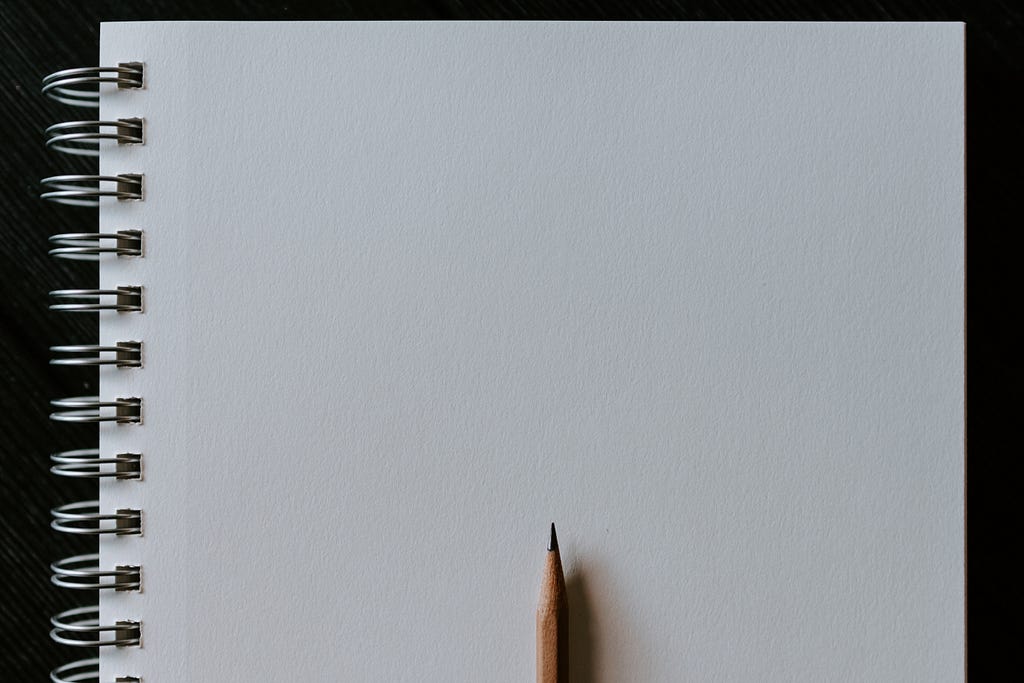 A spiral-bound notebook of white paper open to a blank page, with a sharpened wooden pencil laying atop it.
