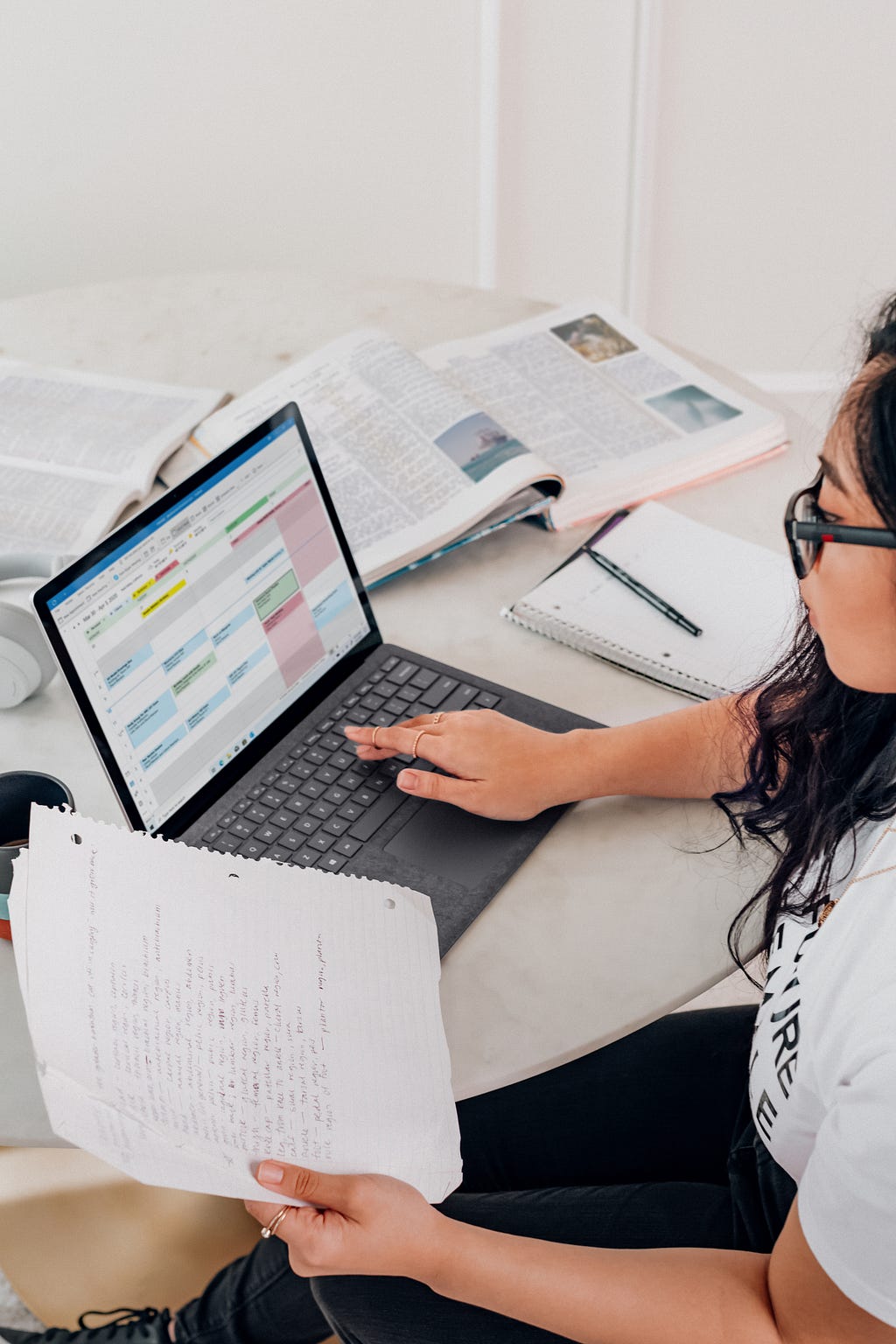 A young woman in glasses looks over a page of handwritten notes, held to the left of her laptop with a calendar display.