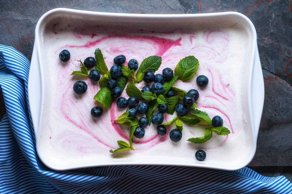 Overhead view of a rectangular ceramic dish with white ice cream with a red swirl & garnished with blueberries + mint leaves.