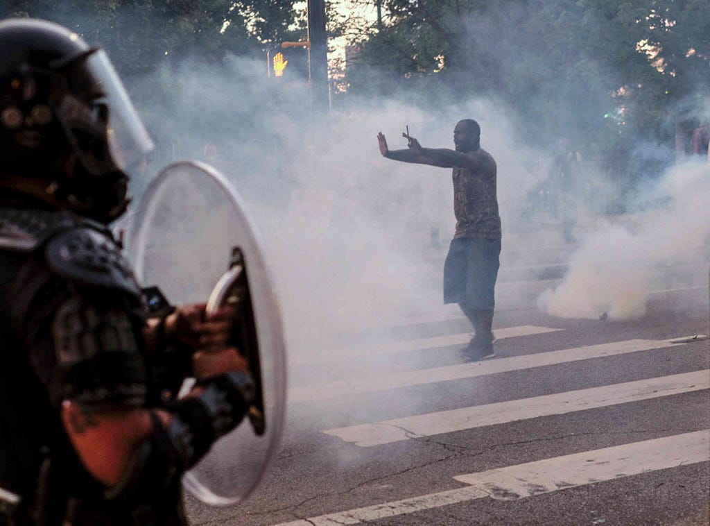 A black protestor stands in the middle of a crosswalk holding his hands up as he is covered in gas. A police officer wearing a helmet stands in frame while holding a shield.