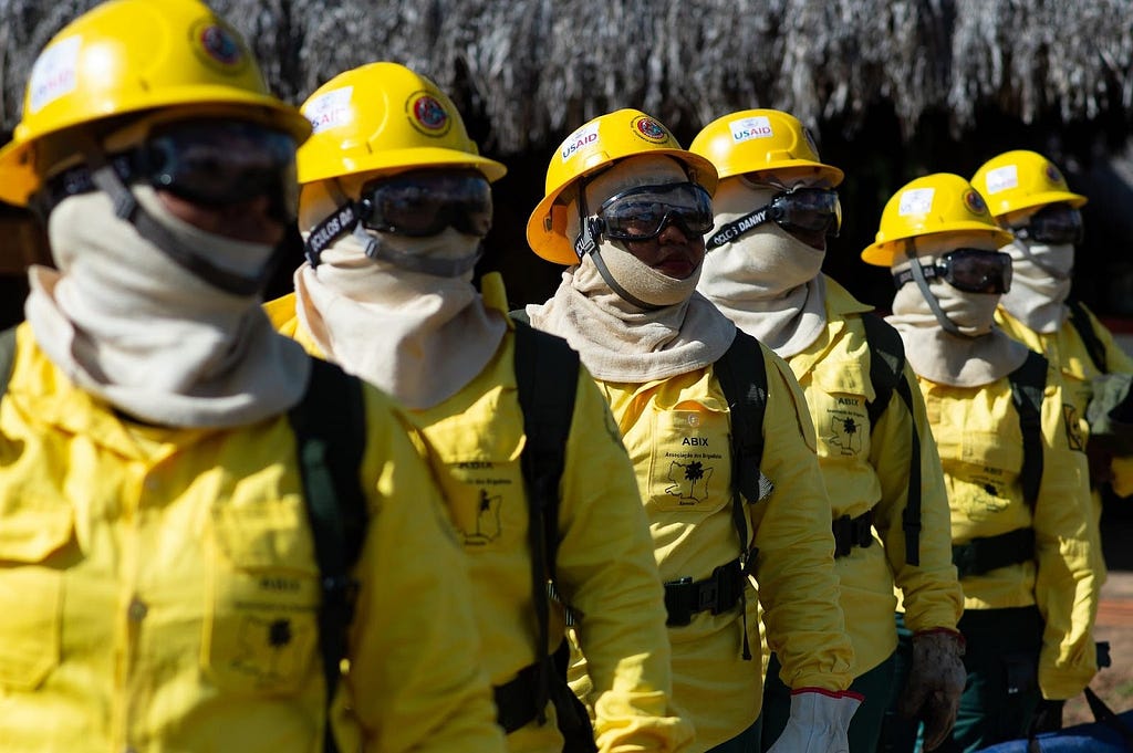 A line of women firefighters dressed in yellow jackets and hardhats, goggles, and off-white colored balaclavas.
