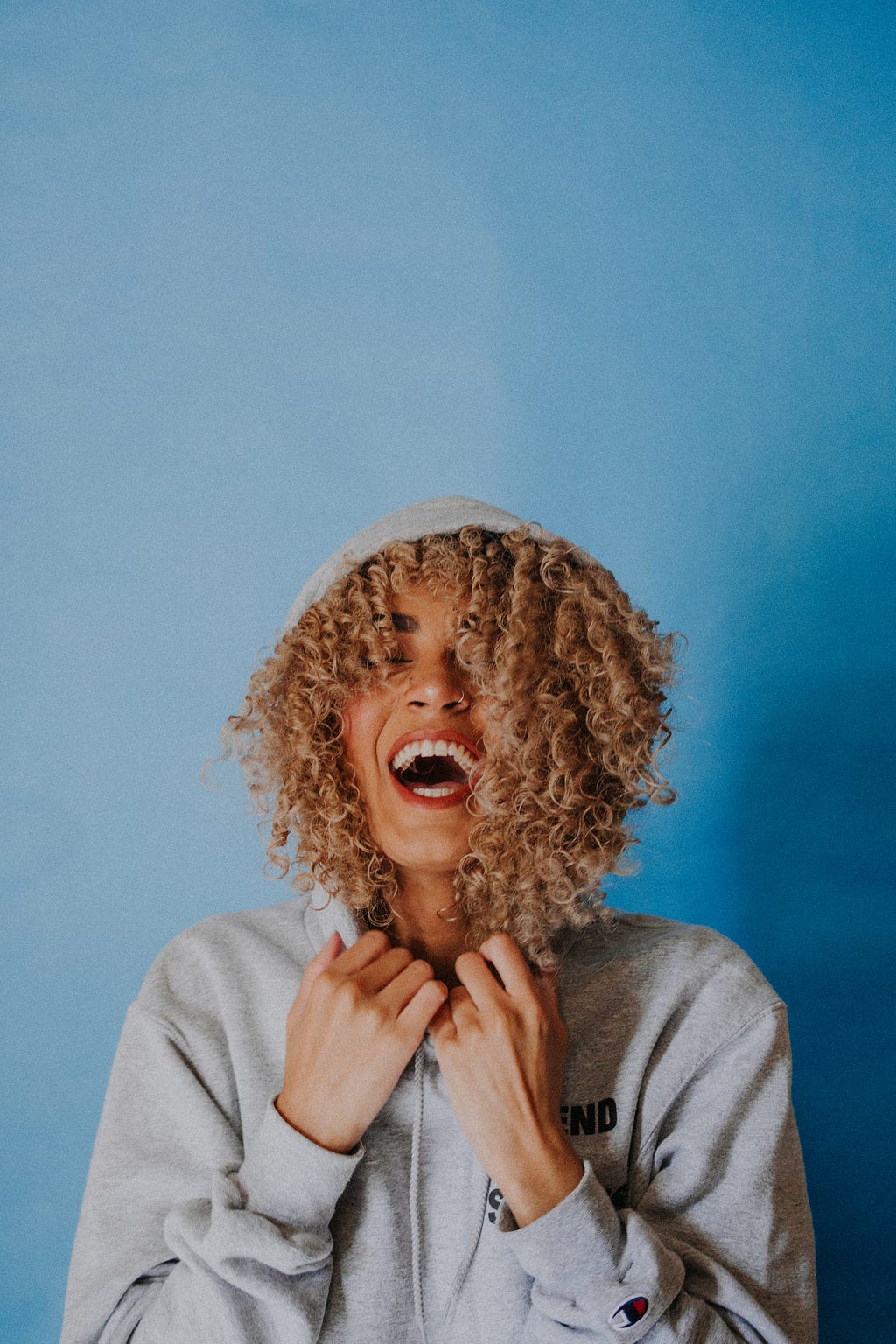 A female of color with curly hair and gray hoody is laughing with her eyes shut in front of a variety of different blues background.