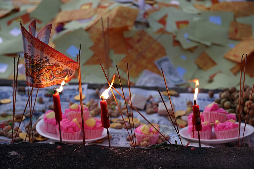 Lit red candles and incense sticks stand amongst an array of traditional food offerings and floating joss paper during the Hungry Ghost Festival, with a vibrant red and gold paper boat symbolizing the journey of ancestral spirits.