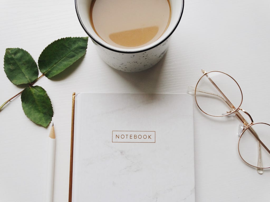 A journal, pencil, glasses & cup of tea on a desk with a green leaf next to it