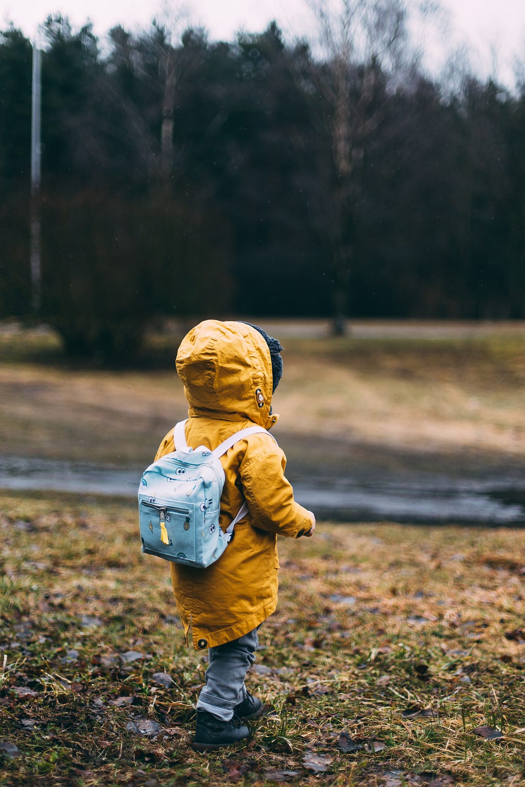A child walking down a hill in early spring, with an orange coat and hood, and a light blue backpack.