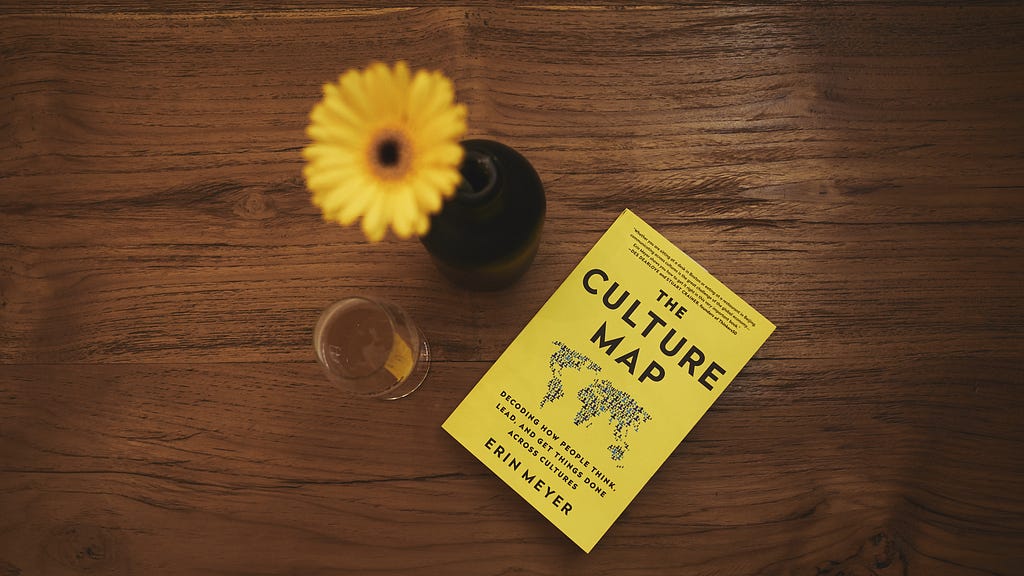 A yellow daisy in vase next to white wine glass and yellow book titled, “The Culture Map” by Erin Meyer on a brown table.