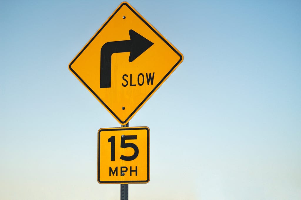 Two road signs stacked on one another indicate a speed limit of 15 miles per hour around a corner. The only copy that accompanies the arrow icon is “Slow” and “15 MPH.”