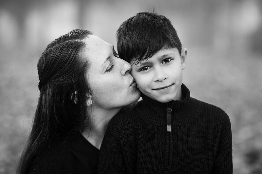 black and white image of mother kissing her son on the cheek in a forest
