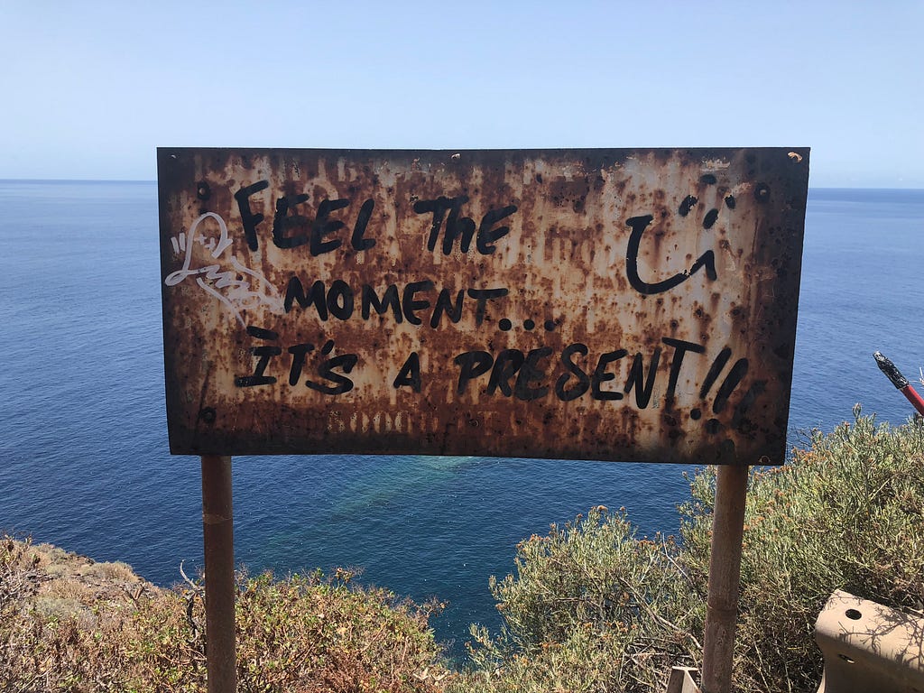 The words “Feel the moment it’s a present” are written on an old sign by the ocean.