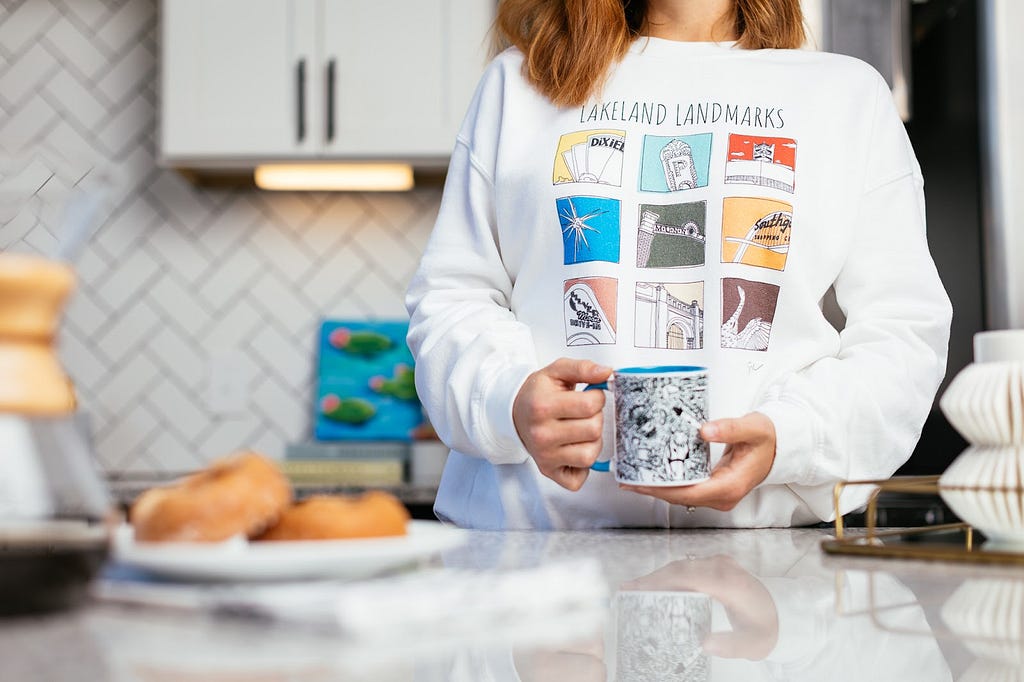 A white woman, visible from the neck down, stands in a kitchen holding a cup of coffee featuring a design by J. Mars Hall while modeling a Lakeland, Florida landmarks sweatshirt. There is a plate of doughnuts and a Chemex coffee maker on the counter in front of her.