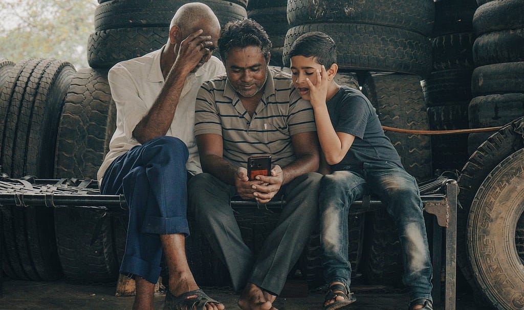 Tres figuras masculinas (un anciano, un adulto y un niño) en un ambiente rodeado por neumáticos, sentados sobre un banco improvisado. Uno de ellos tiene en sus manos un smartphone hacia el cual está dirigida la atención de los tres.