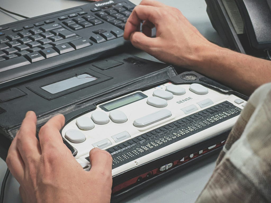 Um teclado braille acoplado em frente à um convencional, aparentemente em uma mesa, sendo operado por duas mãos.