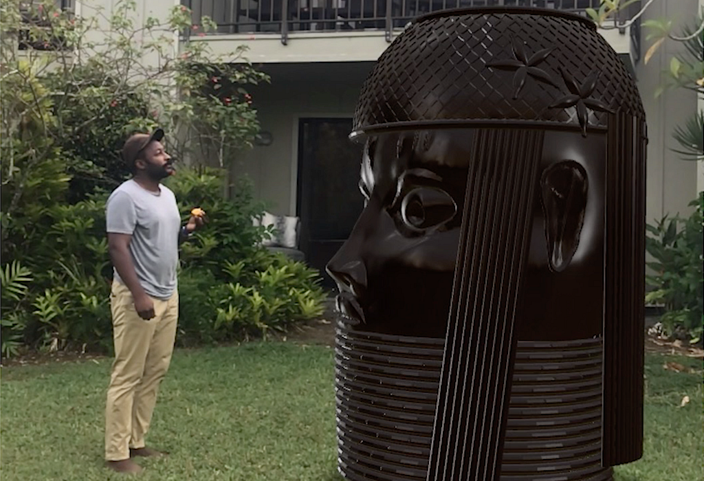 A Black person wearing a baseball cap, grey t-shirt and khaki pants standing on a lawn and gazing at a larger than life-size augmented reality rendering of a bronze King’s Head sculpture from the Kingdom of Benin. The background is the landscaped exterior of an apartment building.
