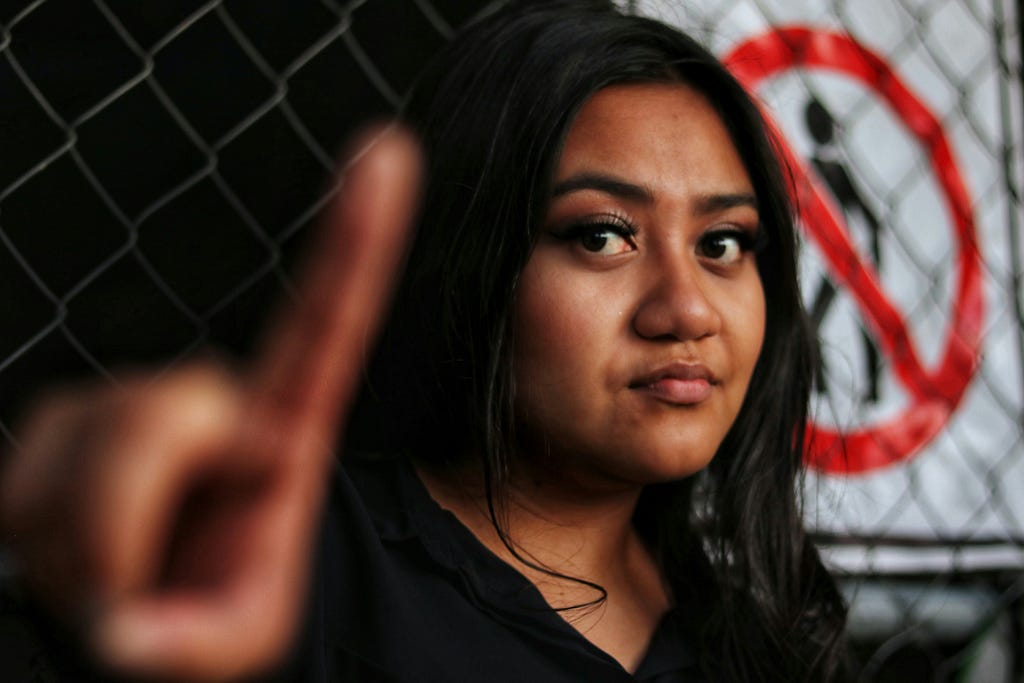 A woman with dark skin and hair holding her finger up as if saying no. Her expression is serious.