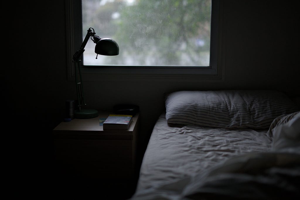 dark shot of a bedroom with one side of the bed empty and a nighttand and book on the left side. the trees and light in the window are lighting up the room
