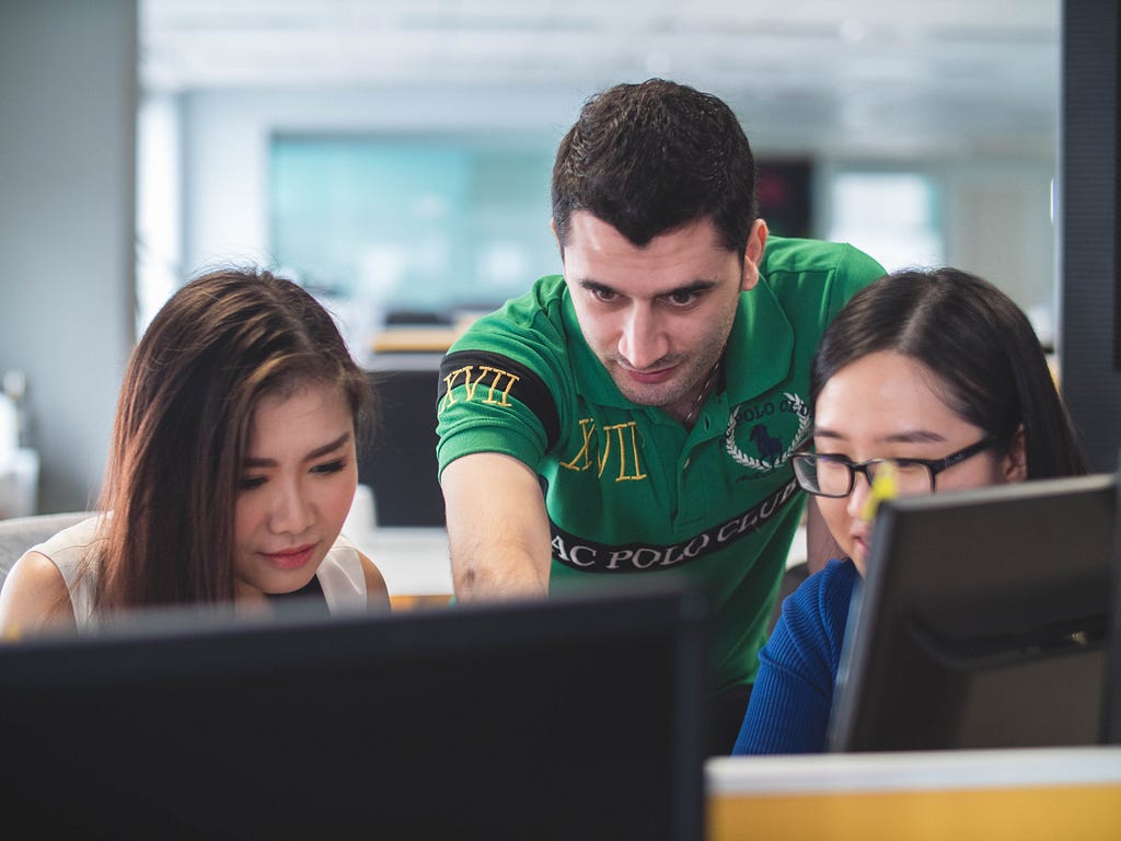 Three people sit at a computer. They point to and discuss what’s on the screen, in the middle of a collaborative discussion.