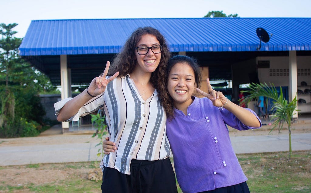 Two women smile and make peace signs in front of a blue-roofed building.