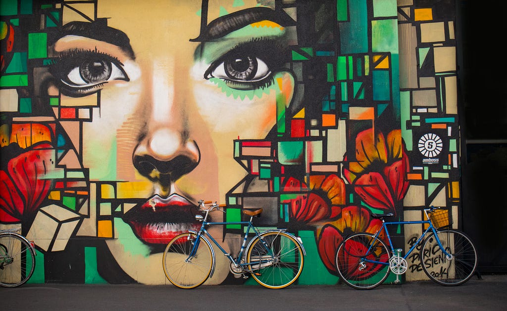 Two bicycles lean against a graffiti wall with a beautiful colourful mural of a woman’s face