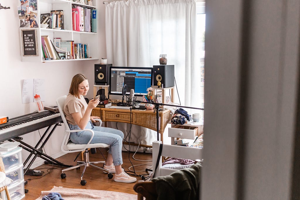 A woman sits at her desk in her bedroom. She is on her phone. On her desk is her computer. Behind her is a keyboard, next to her is recording equipment. To the far right is her bed and on the walls are shelves filled with books.
